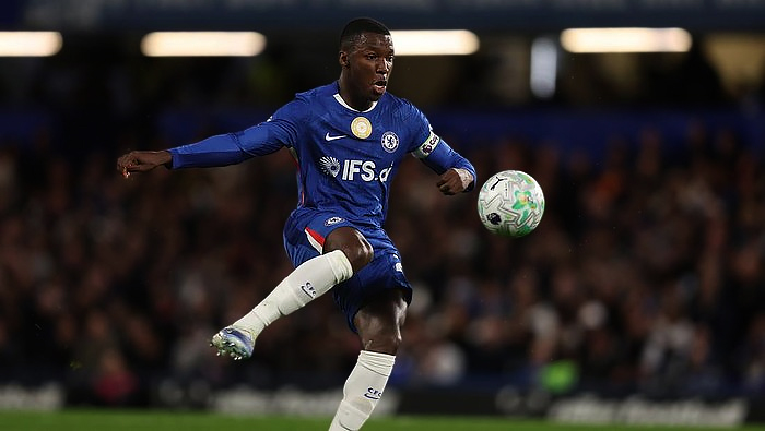 Soccer player in a blue jersey kicks a ball mid-air during a match, stands blurred in the background.