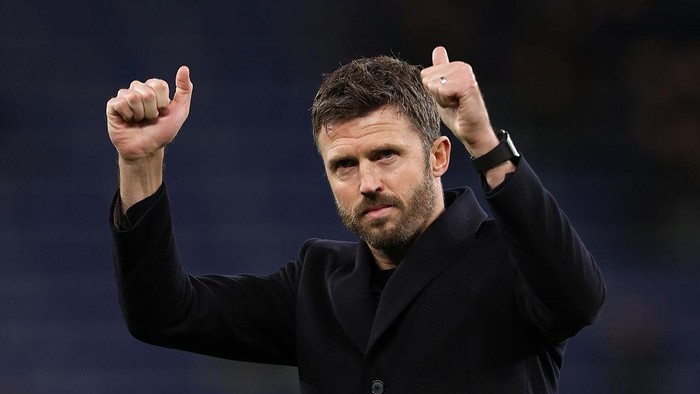 Male coach in a dark coat raising both thumbs in celebration on a football pitch