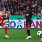 Soccer moment: a red‑and‑white attacker charges the ball as a dark kit defender and orange‑gloved goalkeeper reach for it near the goal; crowd in the stands behind.