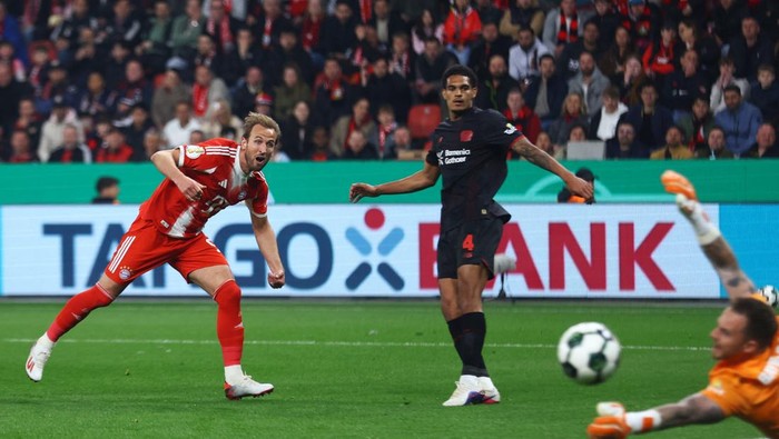 Soccer moment: a red‑and‑white attacker charges the ball as a dark kit defender and orange‑gloved goalkeeper reach for it near the goal; crowd in the stands behind.