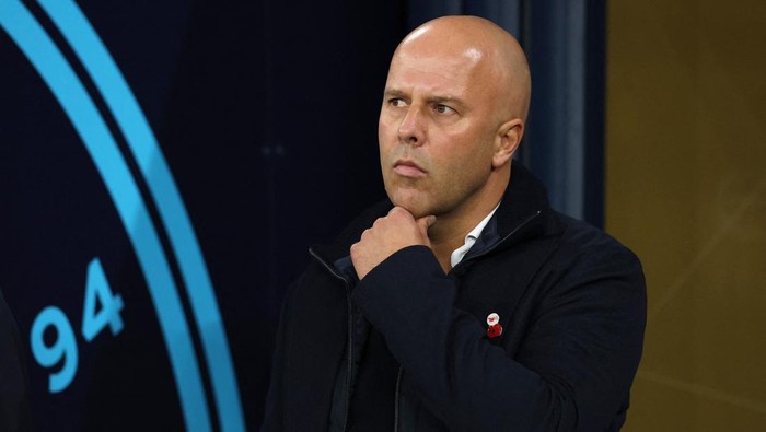Bald man in a dark jacket stands with his hand on his chin, looking thoughtful against a dark background with blue circular markings at the left.