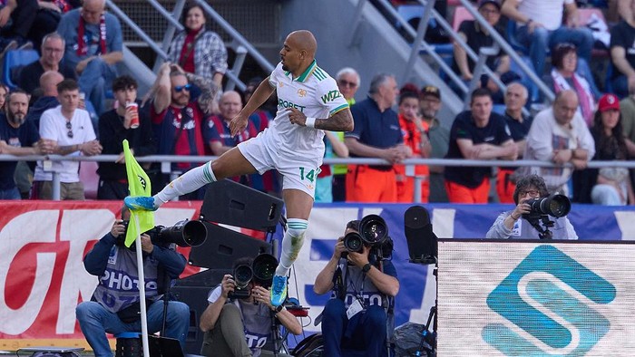 Soccer player in white-green kit jumps near the sideline, kicking a flag while photographers film and spectators watch from the stands.
