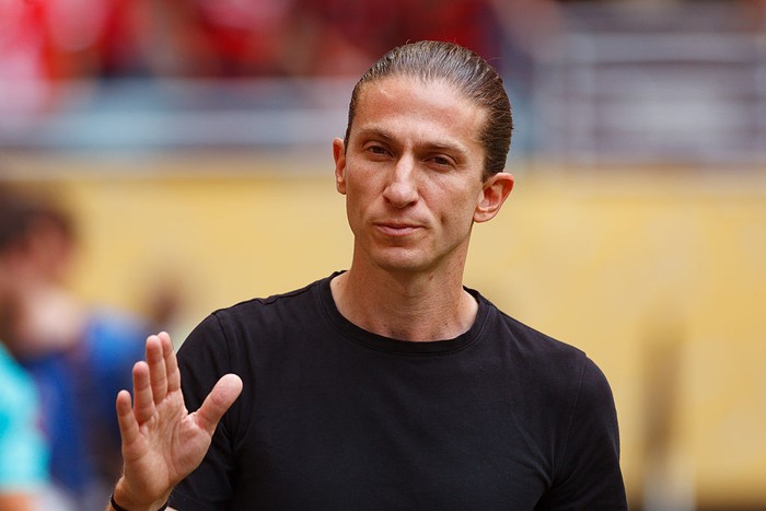 Man with slicked-back hair in a black T-shirt raises his hand in a stop gesture outdoors.
