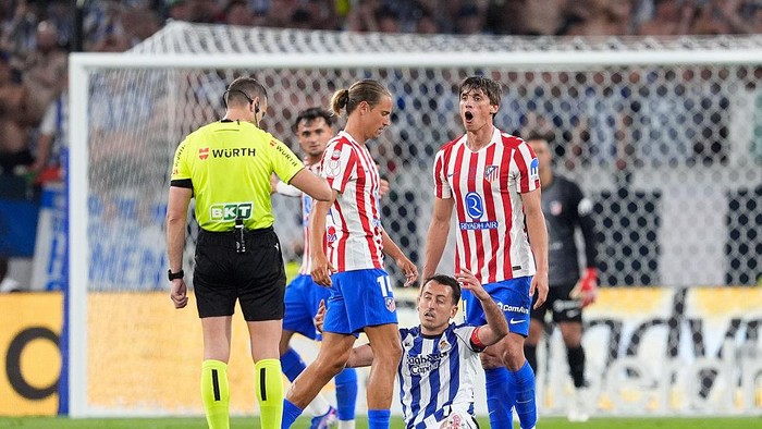 Soccer scene with a neon-yellow referee and players in red-and-white stripes near the goal; one player is on the ground.]","Football match moment near the goal as players in red-and-white attack and a fallen blue-and-white player is attended by teammates.","Referee in bright yellow overseeing a tense moment as players in red-and-white strips cluster near the goal.","Somber moment during a soccer match: a player on the ground while teammates and a referee watch near the goal.","Goal-area action in a soccer match with players in red-and-white stripes and blue uniforms and a bright referee nearby.