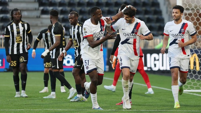 Soccer players celebrate a goal on the field: PSG players in white jerseys with red/blue trim, while opponents in black-and-gold stripes stand in the background.