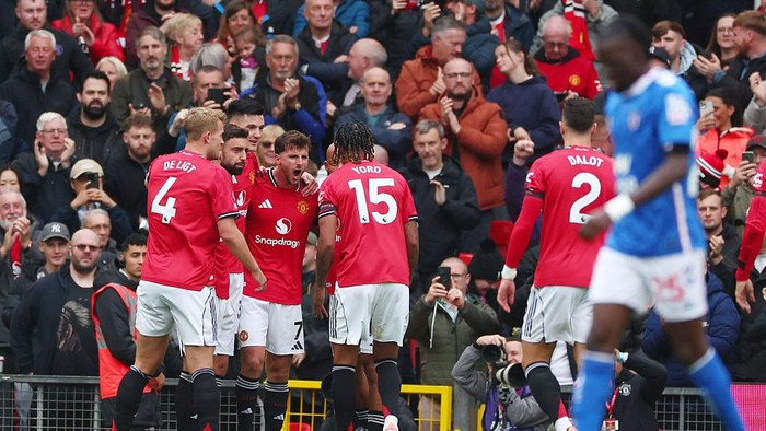 Manchester United players in red celebrate a goal on the pitch as fans applaud in the stands; jersey numbers 4, 15 and 2 are visible.