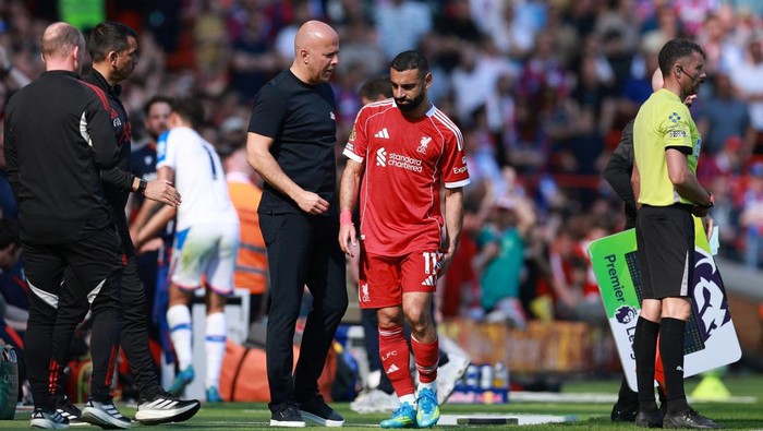 Soccer player in red Liverpool kit walks off the field while a coach speaks to him on the sideline, with officials nearby.
