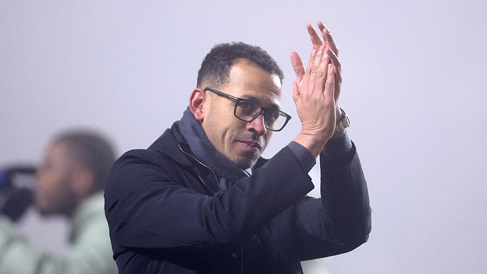 Man wearing glasses and a dark jacket claps his hands in front of a blurred background, focusing on the moment of applause.
