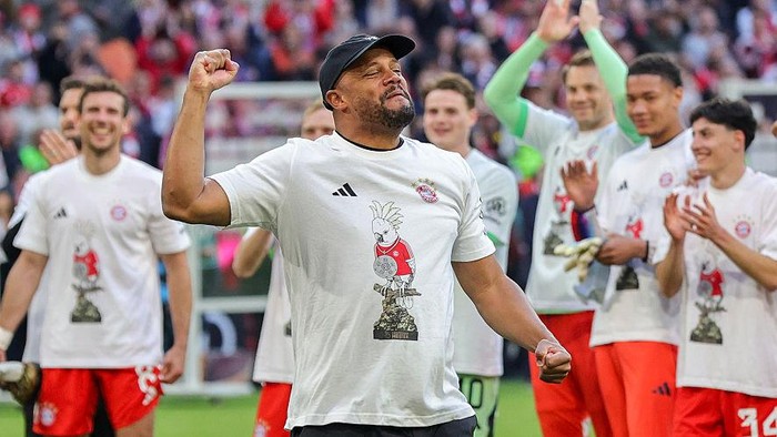 Soccer coach raises a clenched fist in celebration while teammates applaud on a sunny field.