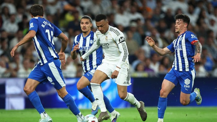 Real Madrid player in white controls the ball while surrounded by four opponents in blue and white striped kits on a green field outside the penalty area.