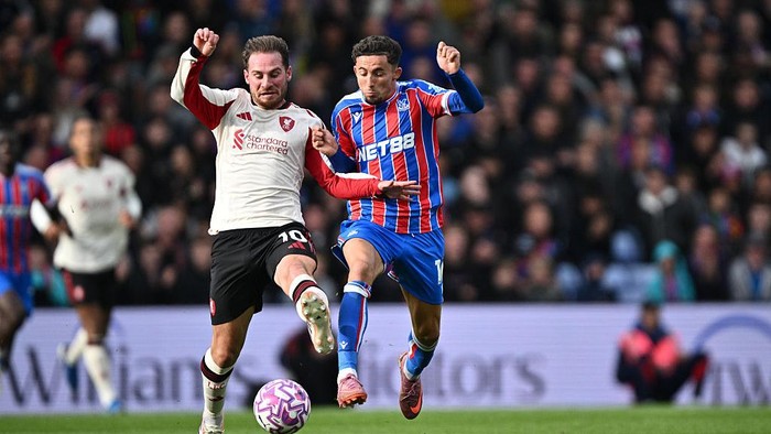Two soccer players duel for the ball during a match, one in a white jersey and the other in blue-and-red stripes, both kicking toward the ball as spectators watch.