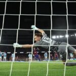 Soccer goalkeeper dives to his right to block a shot near the goal during a nighttime match, with teammates and spectators in the background.