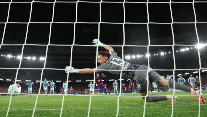 Soccer goalkeeper dives to his right to block a shot near the goal during a nighttime match, with teammates and spectators in the background.
