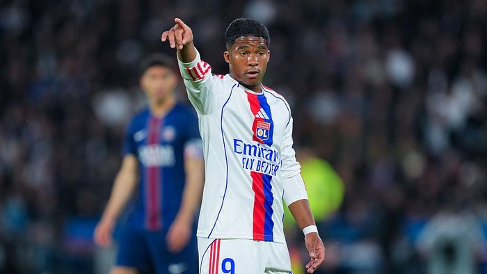 Footballer in a white Olympique Lyonnais kit points toward the field during a match, with an opposing player blurred in the background.