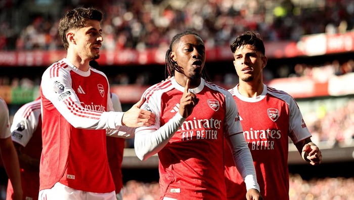 Three Arsenal players in red and white jerseys celebrate on the pitch after a goal, with one in mid-shout and two teammates beside him.