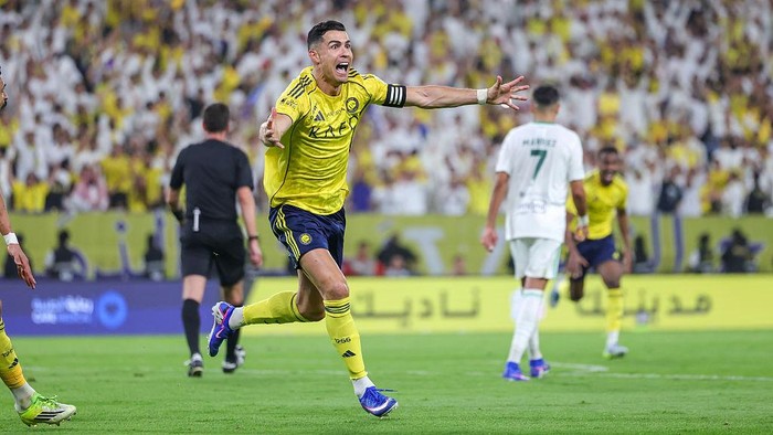 Soccer player in a yellow kit with a captain’s armband celebrates a goal on the green field as fans cheer in the stands.