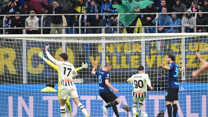 Soccer players in white and blue-black kits clash near the goal as fans watch from the stands behind the net.