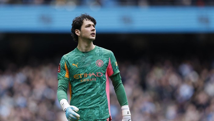 Soccer goalie in a green kit with gloves stands on the pitch as blurred fans watch from the stands behind him.