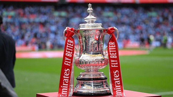 Silver trophy on a red pedestal with red ribbons reading 'Emirates Fly Better' in a stadium setting.
