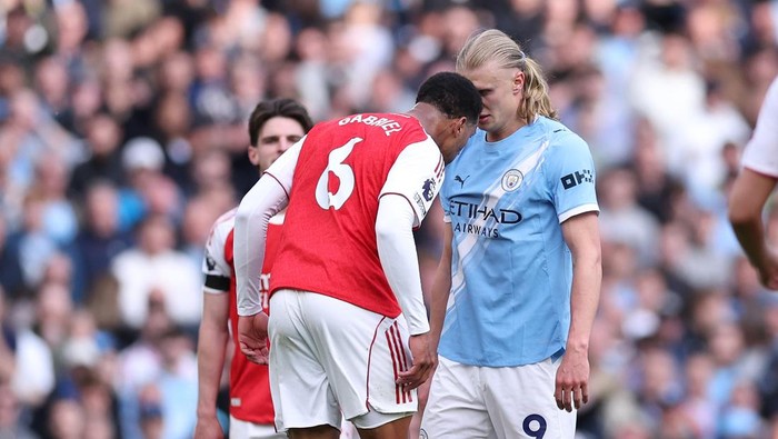 Two soccer players from Arsenal (red jersey with number 6) and Manchester City (light blue jersey) lean in toward each other during a match, heads close in a confrontational moment.