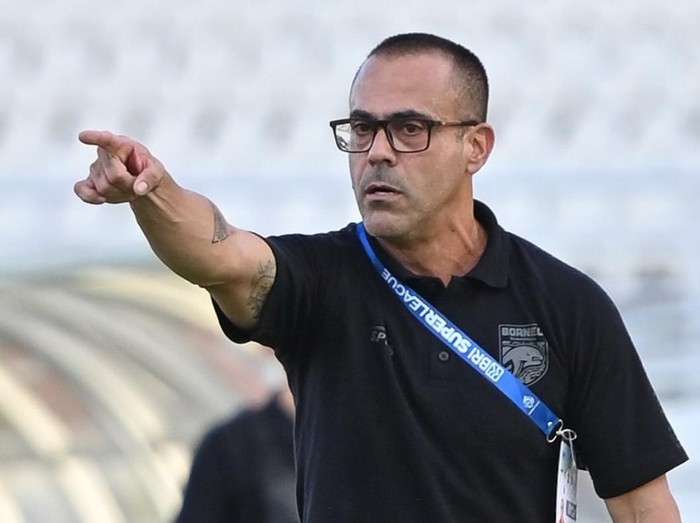 Coach wearing a black polo and glasses points emphatically toward the field from the sideline.