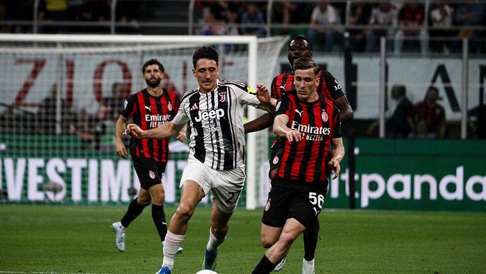Juventus player in black-and-white kit dribbles the ball as Milan players in red-and-black chase near the goal.