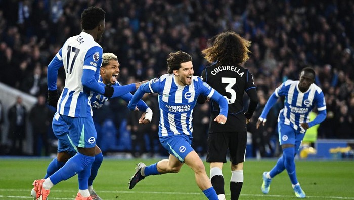 Brighton players in blue-and-white striped kits celebrate on the field after a goal, with Cucurella (number 3) in black defending nearby.