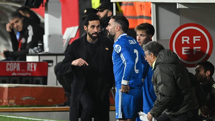 Two men converse on the soccer sideline: one in a black coat, the other in a blue jersey, with teammates nearby and an RF EF sign in the background.