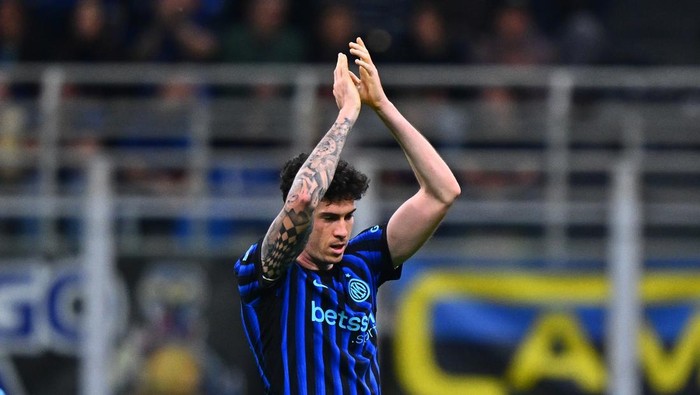 Soccer player in blue and black striped jersey clapping on the field with visible arm tattoos.In the background stadium seats blur.