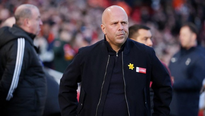 Bald man in a black zip jacket with a yellow flower pin, standing on a football sideline with a focused expression and a blurred crowd behind him.