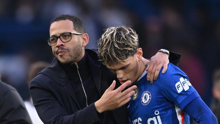 Two men, one in glasses and a dark jacket, supports a Chelsea player by placing an arm around his shoulders after a game.