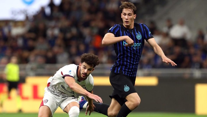 Two soccer players compete for the ball during a match, one in a blue-and-black striped jersey and the other in white, with a stadium crowd in the background.