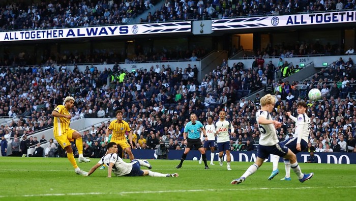 Soccer match action: players chase the ball as a defender slides on the ground and a yellow-clad opponent stands nearby in a packed stadium.