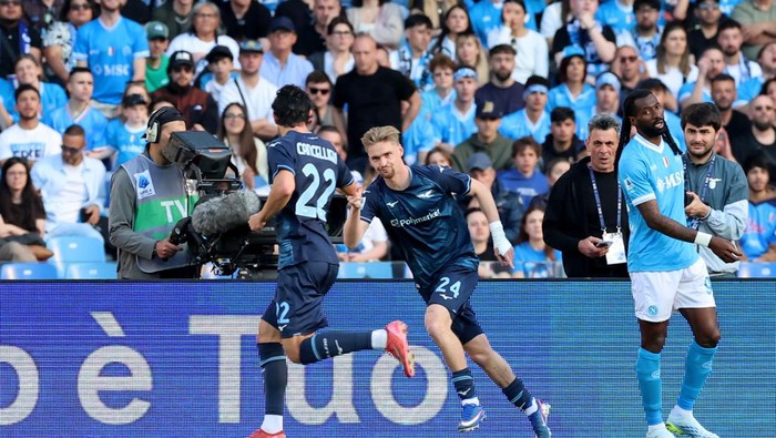 Soccer players in blue celebrate a goal on the field as a cameraman films and a crowded stand watches in the background, near ad boards.