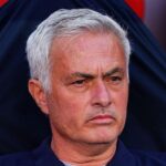 Middle-aged man with short gray hair and a serious expression sits in a dark jacket against a red background.