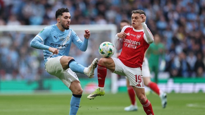 Two soccer players from Manchester City and Arsenal compete for the ball mid-kick on a green field with spectators in the background.