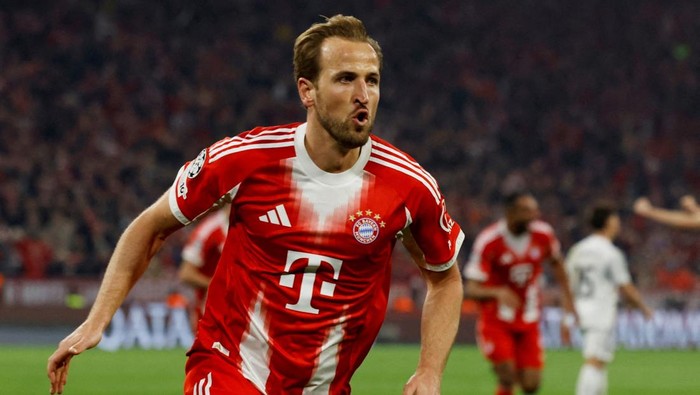 Soccer player in a red Bayern Munich kit sprinting on the field during a match, mouth open in a shout, stadium crowd behind him.