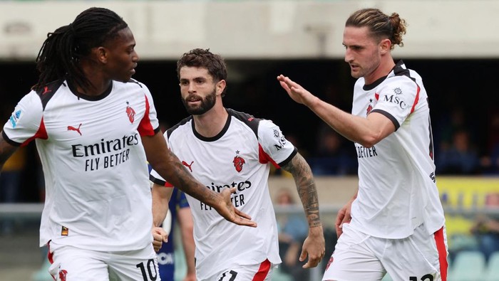 Three soccer players in white jerseys celebrate on the field, one reaching out as another smiles.