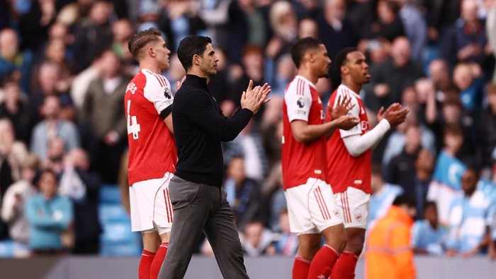 Arsenal players in red and white kits applaud on the field as a coach in black claps, with a crowd in the background.