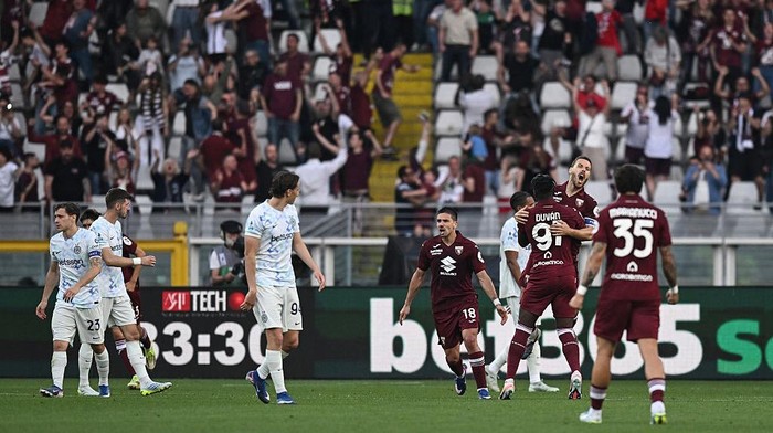 Soccer players on a field celebrate a goal as maroon and light-blue teams clash, with cheering fans in the stands behind.