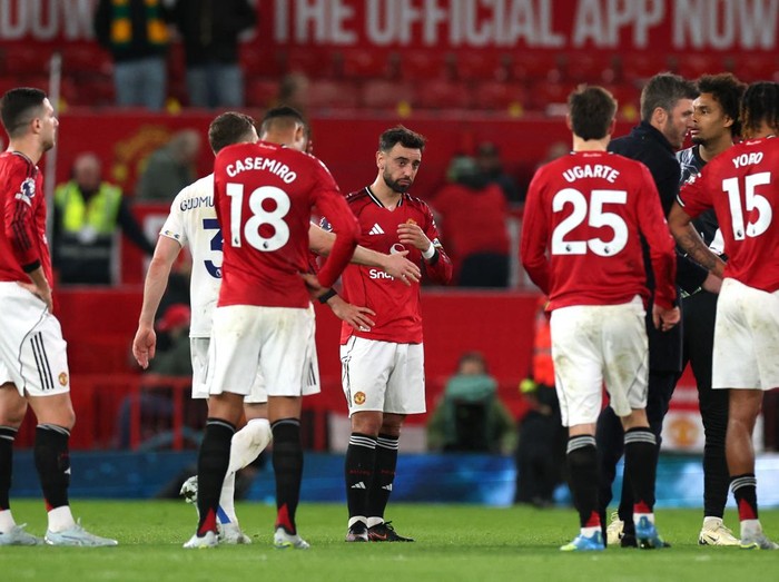Manchester United players wearing red jerseys stand in a group on the pitch, central player looking down with hands at chest.