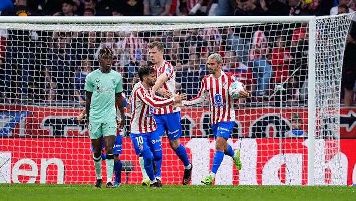 Soccer players in red-and-white striped jerseys celebrate near the goal as a teammate holds a ball by the net.