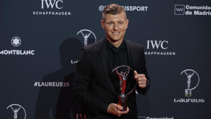 Man in a black suit smiles while holding a Laureus Award trophy against a dark backdrop with sponsor logos.