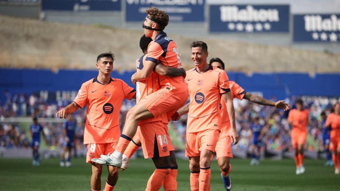 Soccer players in orange kits celebrate a goal on the field, two teammates hug mid-air as others cheer in the background on a sunny day.