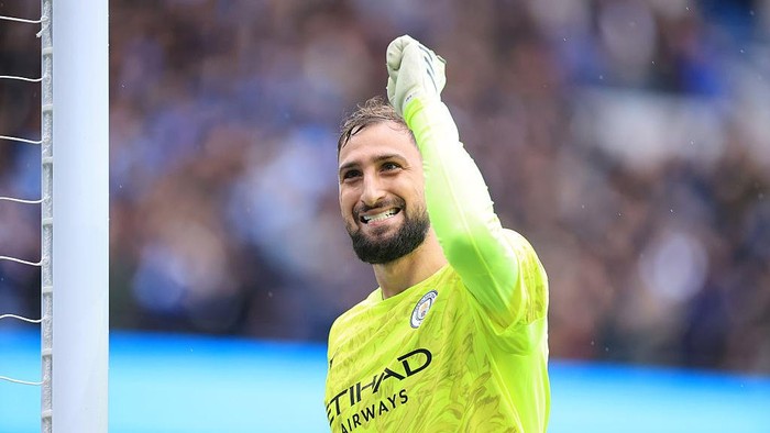 Manchester City goalkeeper in a neon-yellow kit raises a fist in celebration beside the goal post, smiling at the crowd.