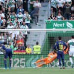 Soccer match scene: goalkeeper in orange dive saves near a white goal with players in white and blue nearby and a crowded stadium behind.