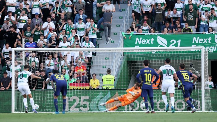 Soccer match scene: goalkeeper in orange dive saves near a white goal with players in white and blue nearby and a crowded stadium behind.