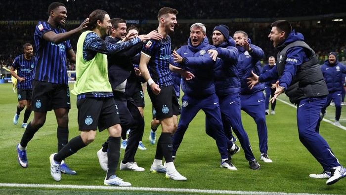 Soccer players in blue-and-black striped jerseys celebrate on a grassy pitch, high-fiving and cheering with coaches nearby during a match.
