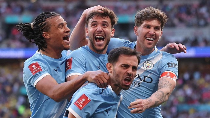 Manchester City players celebrate a goal, embracing each other on the pitch in light blue kits.