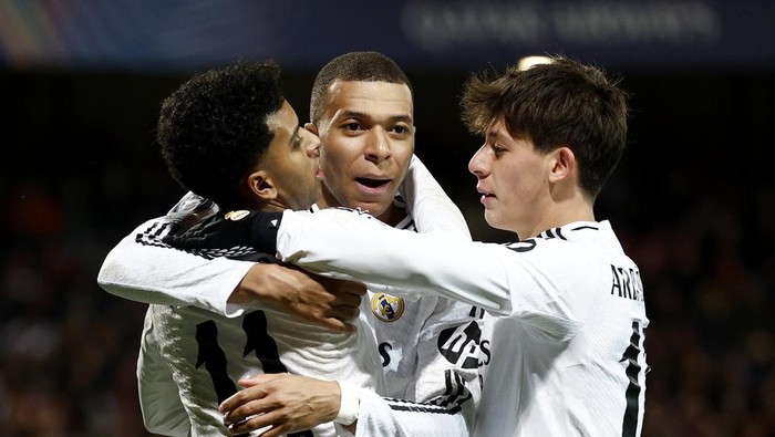 Three soccer players in white uniforms celebrate with a group hug on the field after a goal.